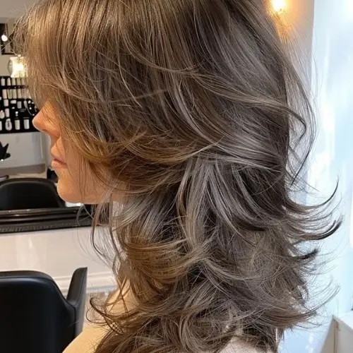 Side profile of a woman with a feathered wolf cut featuring soft face-framing layers, wispy curtain bangs, voluminous textured layers, flipped-out ends, and an ash brown hair color styled with a smooth, bouncy finish.