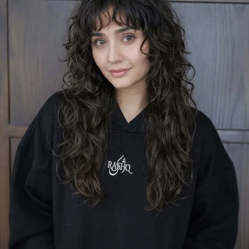 Woman with a long, curly emo wolf cut with wispy bangs.
